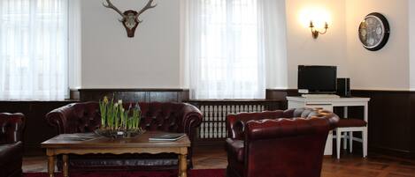 A living room with a wooden coffee table, leather armchairs, a red rug, and a mounted deer head on the wall.