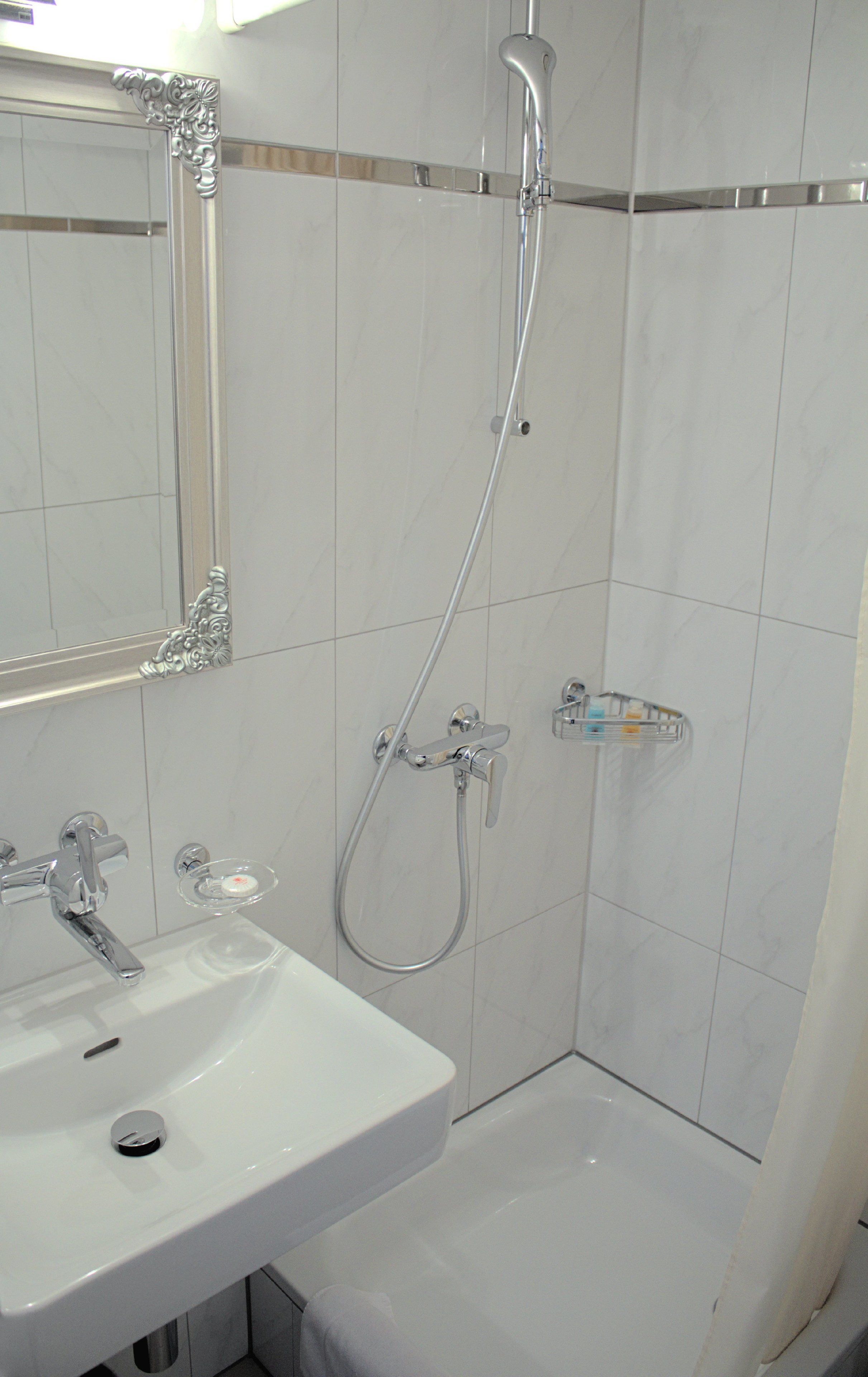 A bathroom with a white sink, a mirror with ornate detailing, a shower with a handheld showerhead, and a glass soap dish.