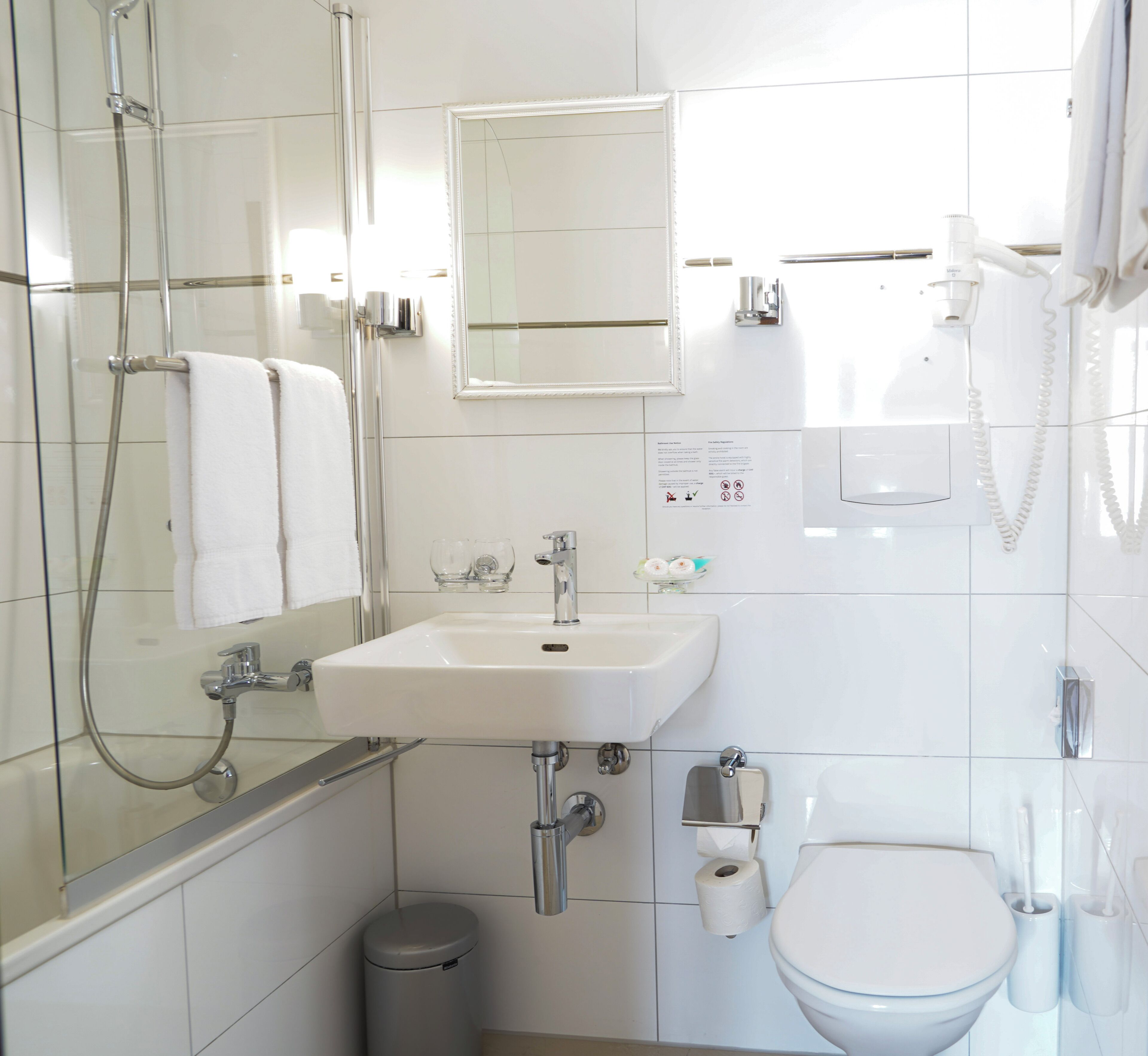 Un baño moderno con ducha de vidrio, un lavamanos con espejo arriba, un inodoro con ducha higiénica y un cubo de basura.