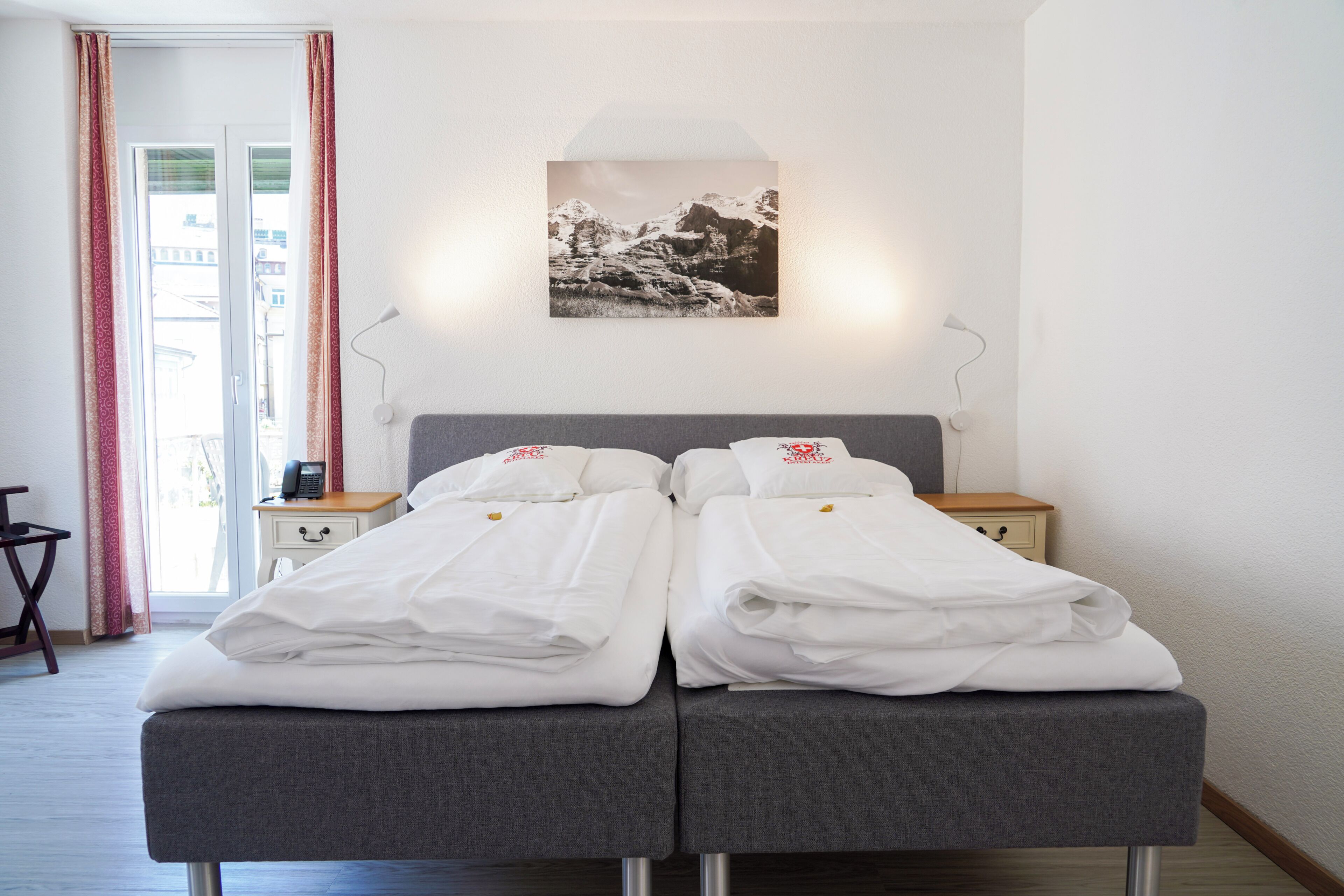 A hotel room with a double bed, bedside tables, a lamp, a painting on the wall, and a view of the outside.
