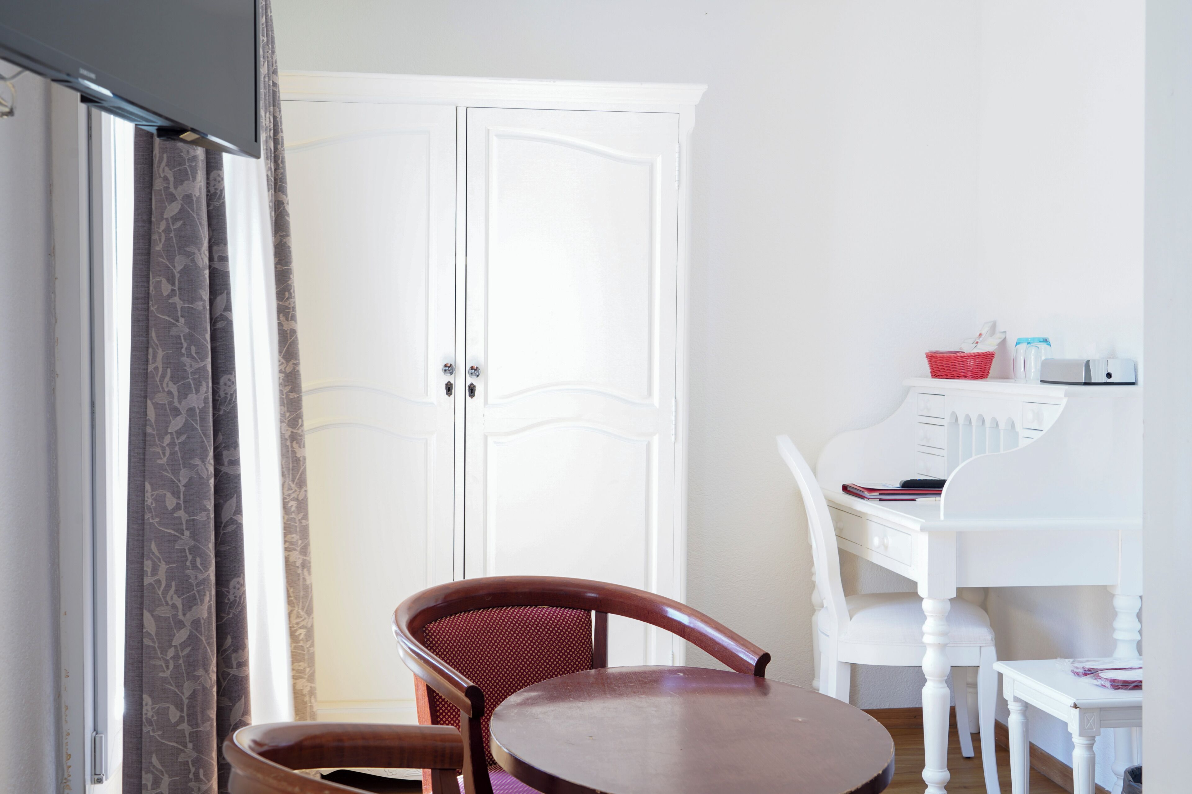 A room with a white wardrobe, a small round table, a wooden chair, and a white desk with a chair.
