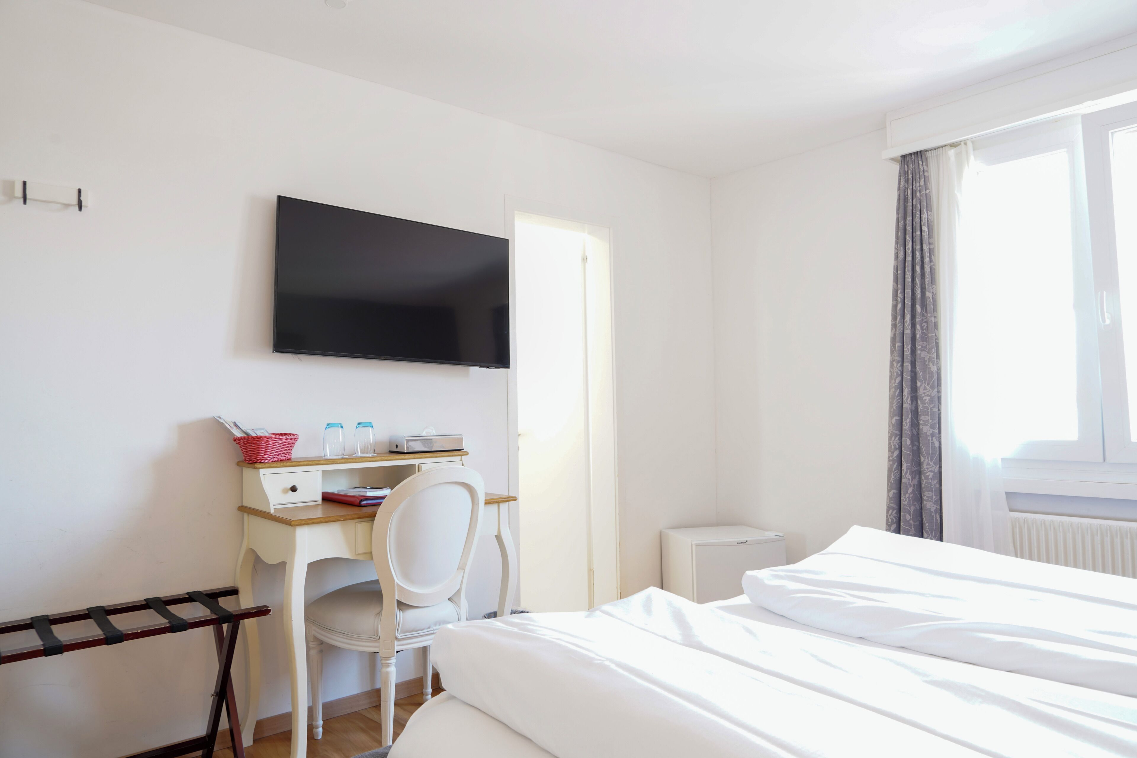 A hotel room with a bed, a desk with a chair, a television mounted on the wall, and a window with curtains.