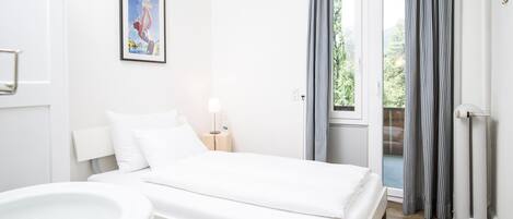 A modern hotel room with a bed, a bedside table with a lamp, a window with curtains, and a view of greenery outside.