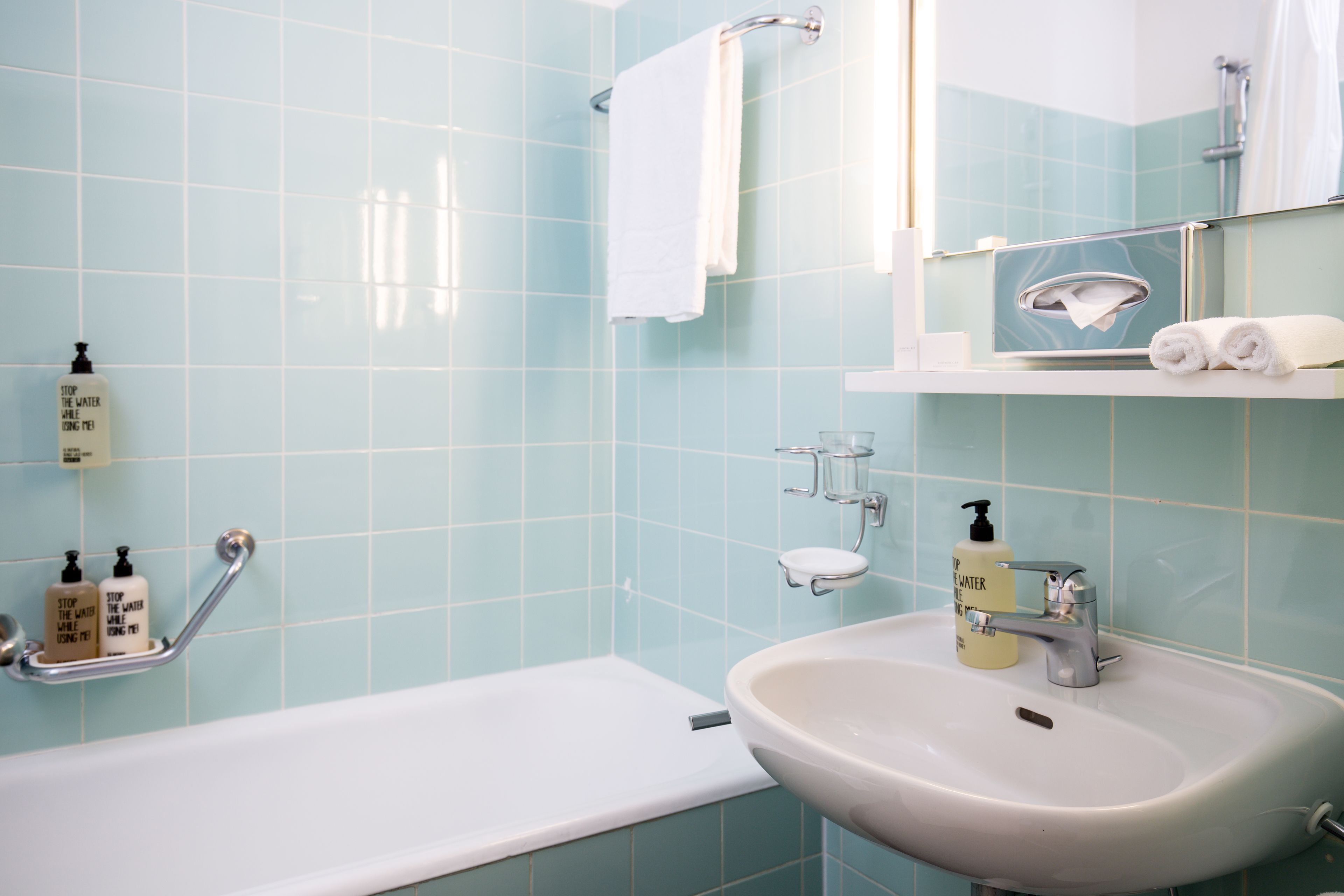 Baño con bañera, lavamanos y espejo. Hay artículos de aseo en la pared y un perchero para toallas.