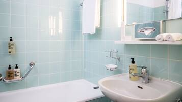 Baño con bañera, lavamanos y espejo. Hay artículos de aseo en la pared y un perchero para toallas.