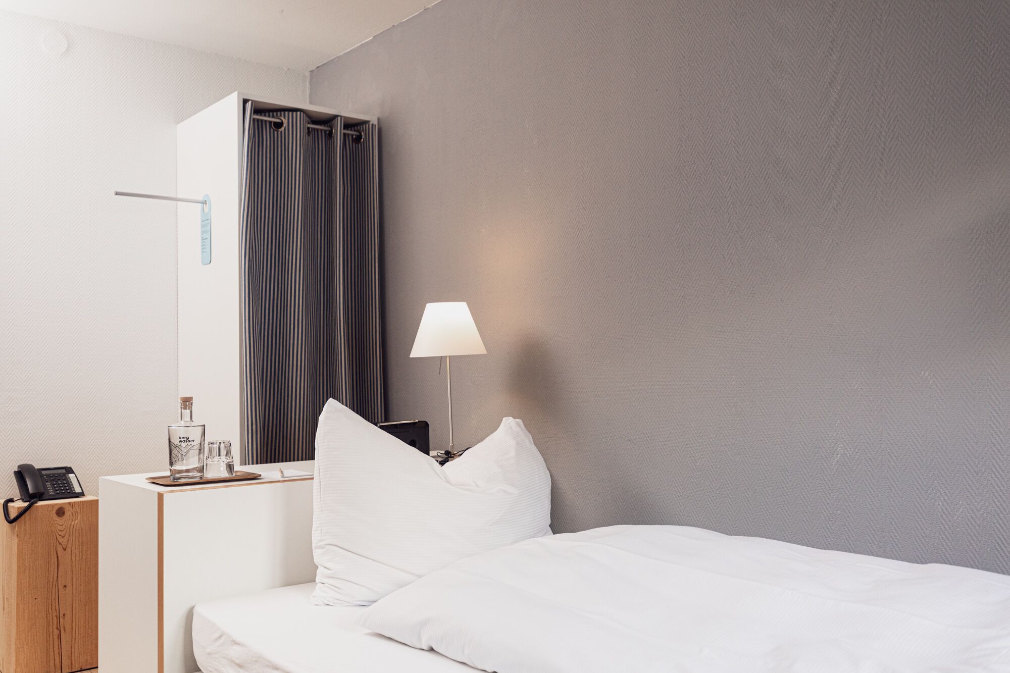 Una camera d'albergo con un letto, un comodino con una lampada, un telefono e un minibar.