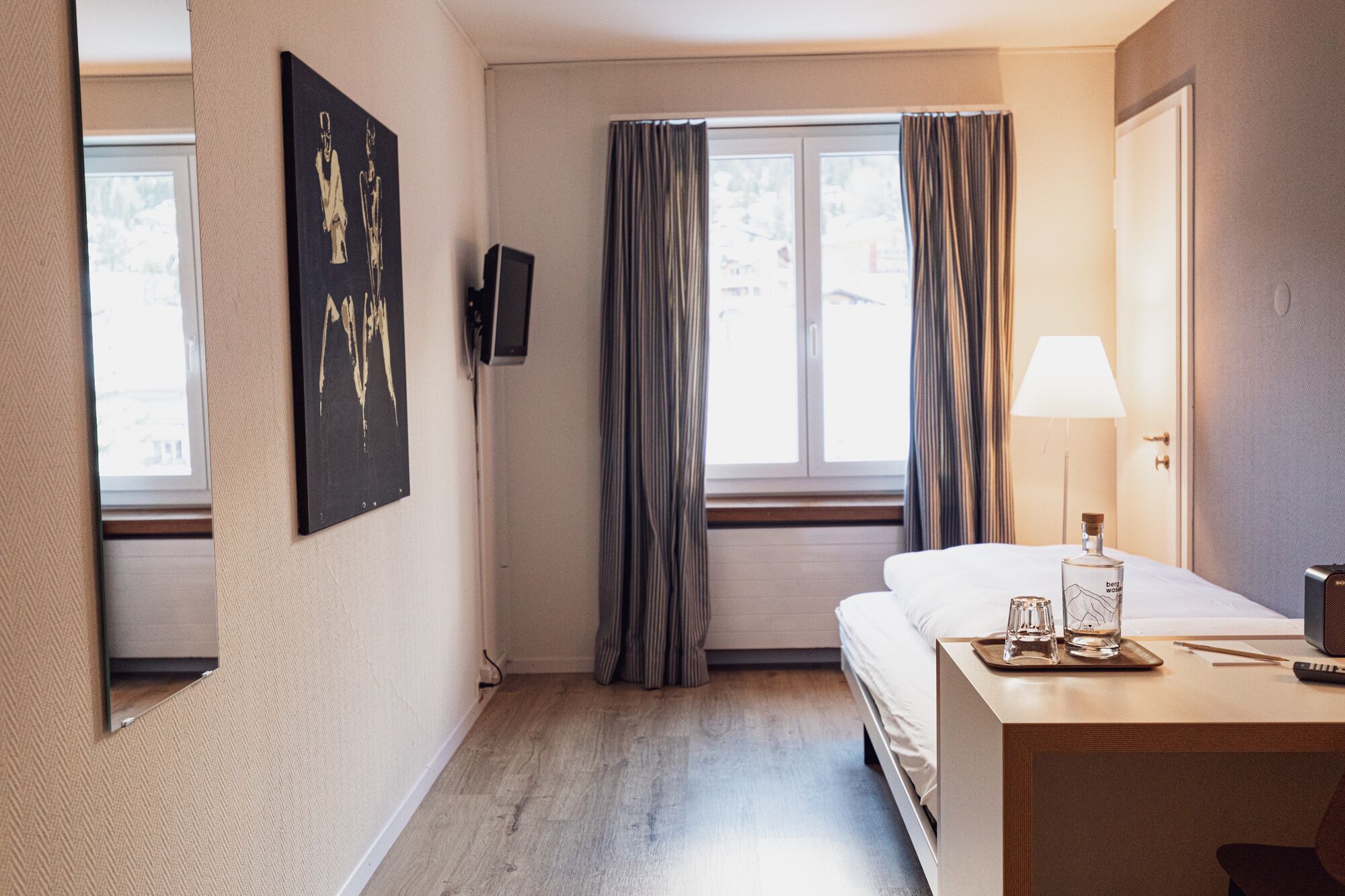 Ett hotellrum med en säng, ett nattduksbord, en lampa och ett fönster med gardiner.
