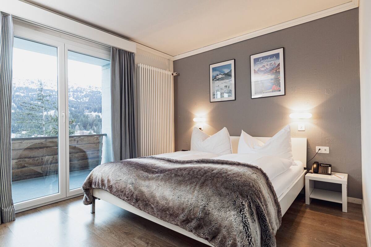 A modern bedroom with a large bed, a nightstand, and a view of a snowy landscape through a sliding glass door.