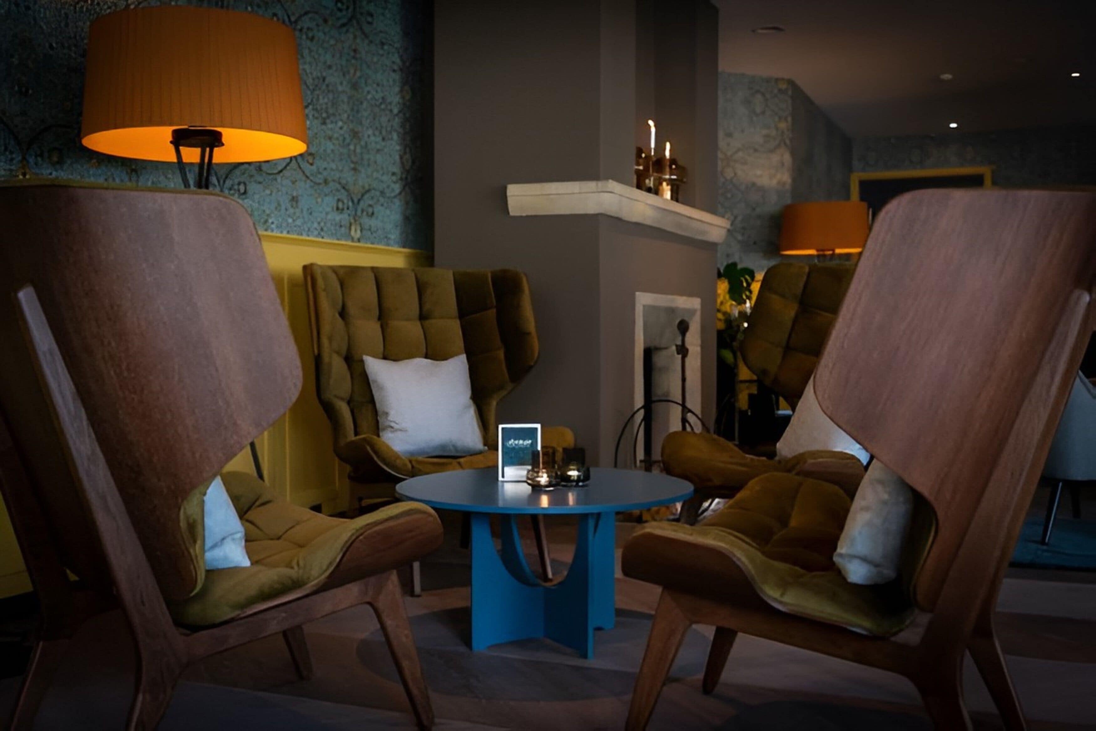 A room with four brown upholstered chairs around a small blue table, a lamp, and a shelf with candles.