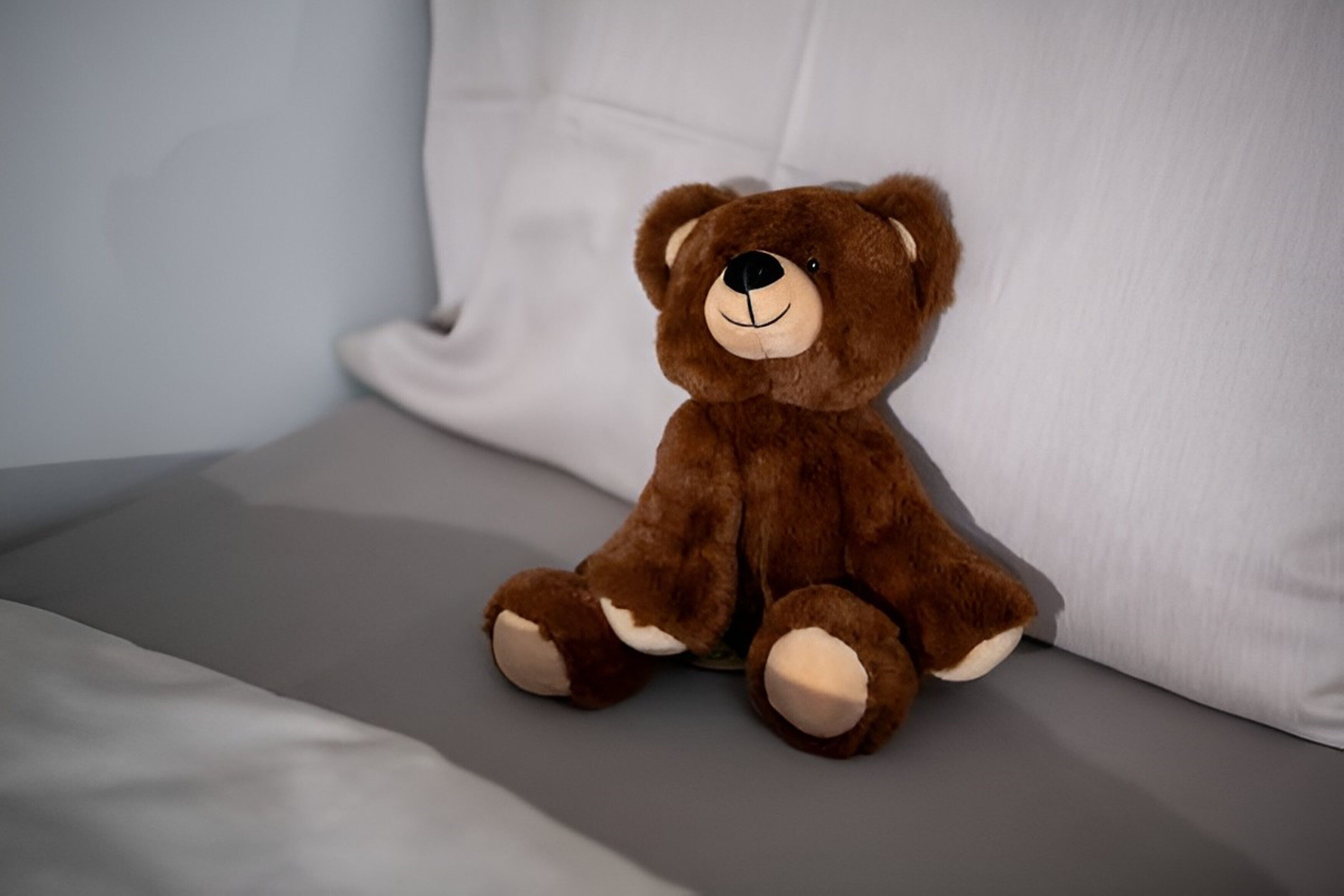 Un osito de peluche marrón sentado sobre una cama con un almohadón blanco.
