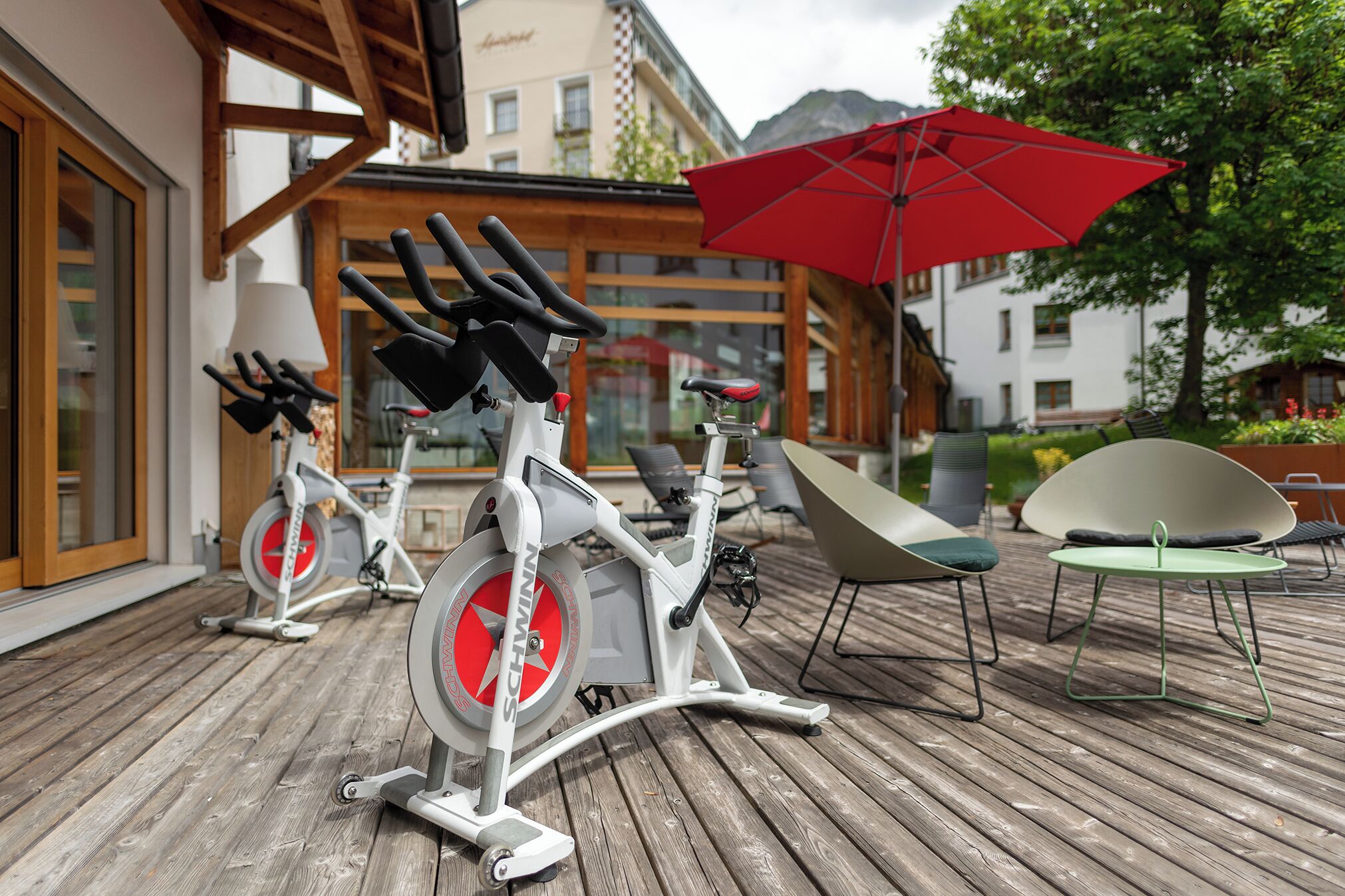 Dos bicicletas estáticas sobre una terraza de madera, una sombrilla roja y mobiliario exterior con vistas a edificios y árboles.
