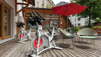 Dos bicicletas estáticas sobre una terraza de madera, una sombrilla roja y mobiliario exterior con vistas a edificios y árboles.