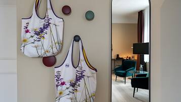 A room with a mirror reflecting a seating area, a wall-mounted bag rack with bags, and a small table with a lamp.