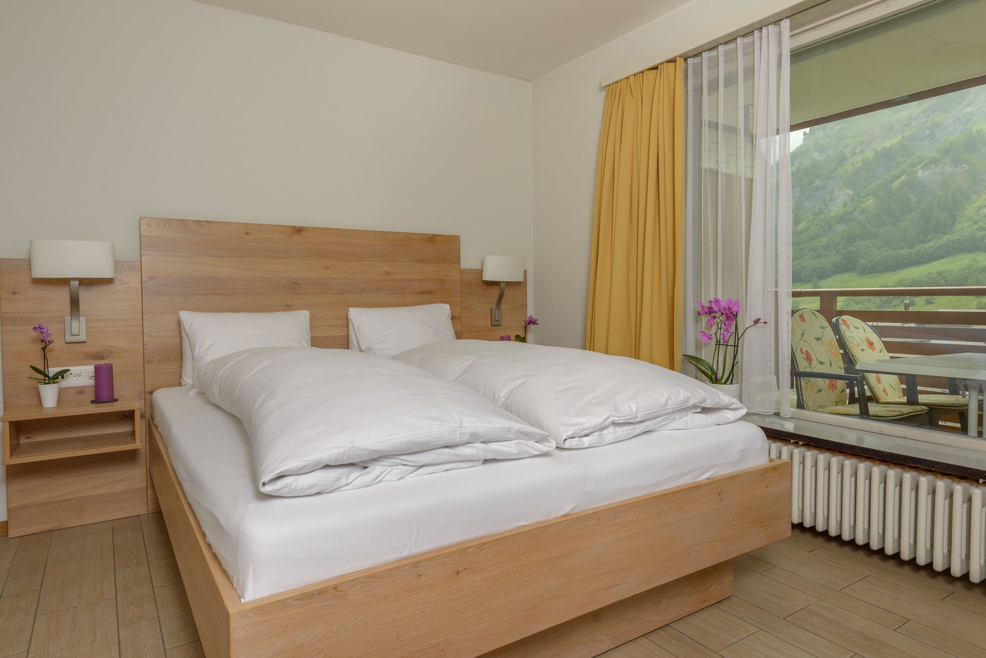 A neatly made double bed with a wooden headboard, bedside lamps, and a view of a balcony with chairs.