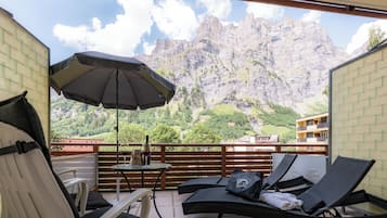 Un balcón con mobiliario exterior, una mesa con una botella y una copa, y vistas a montañas.