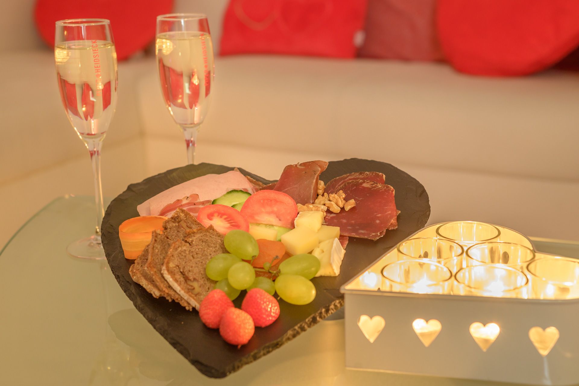 A table with a fruit and cheese platter, two glasses of white wine, and a candle with heart-shaped lights.