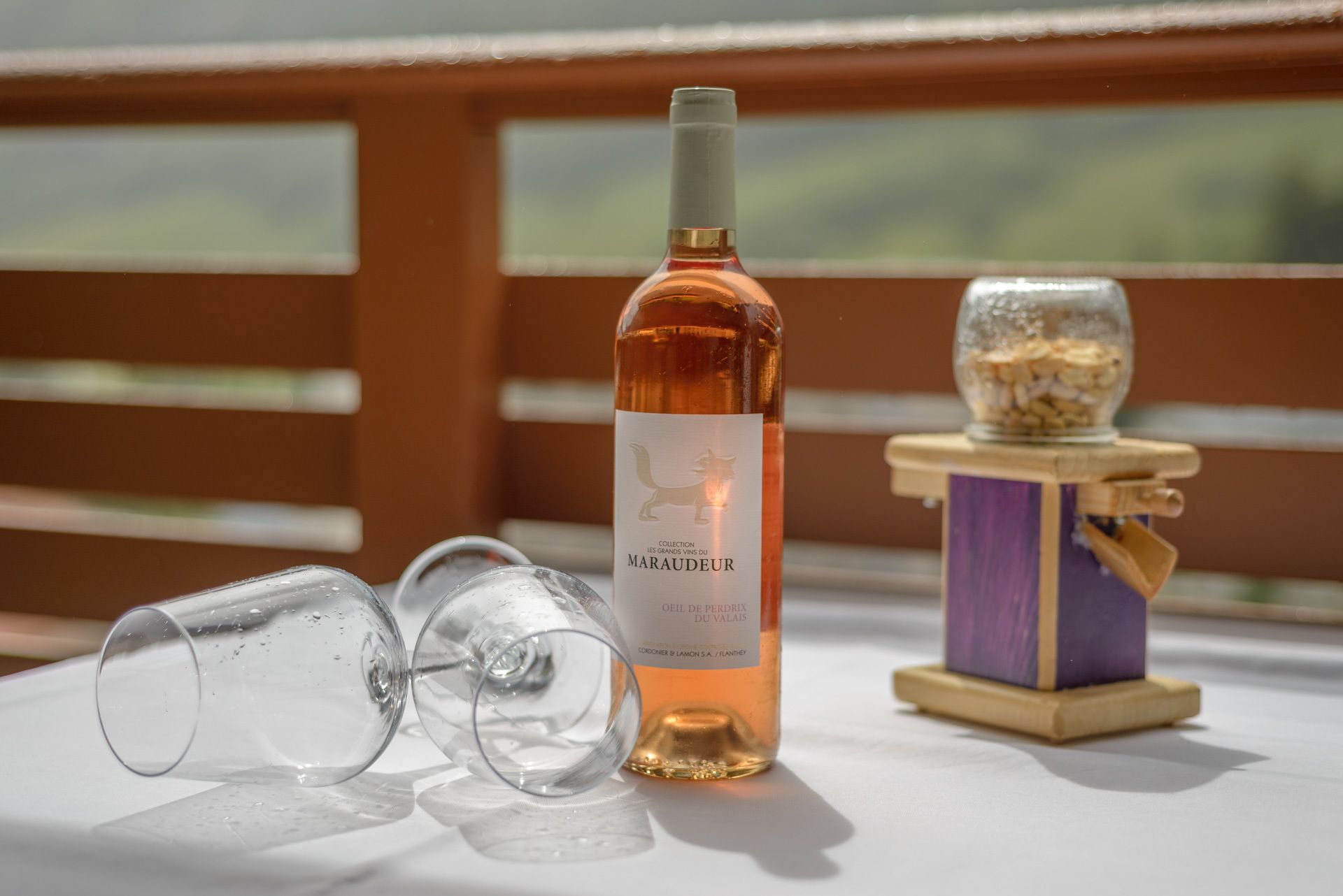 A bottle of wine, empty glasses, and a small container on a table with a view in the background.