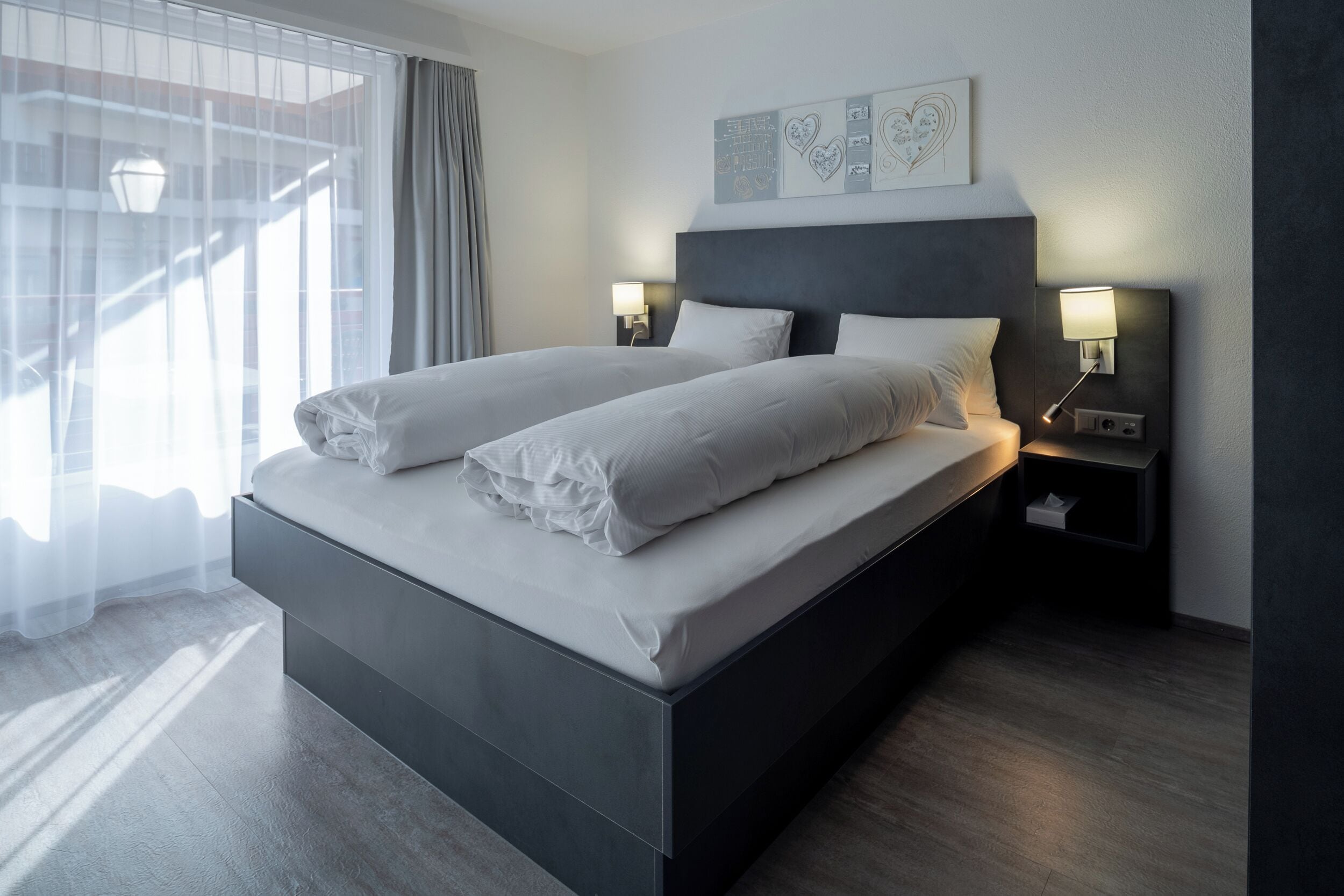 A modern hotel room with a large bed, two bedside tables with lamps, a window with curtains, and a wall with artwork.