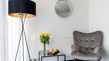 Ein Raum mit einer schwarzen Stehlampe, einem grau gepolsterten Sessel, einem kleinen Tisch mit einer Vase voller gelber Blumen und einer Obstschale.