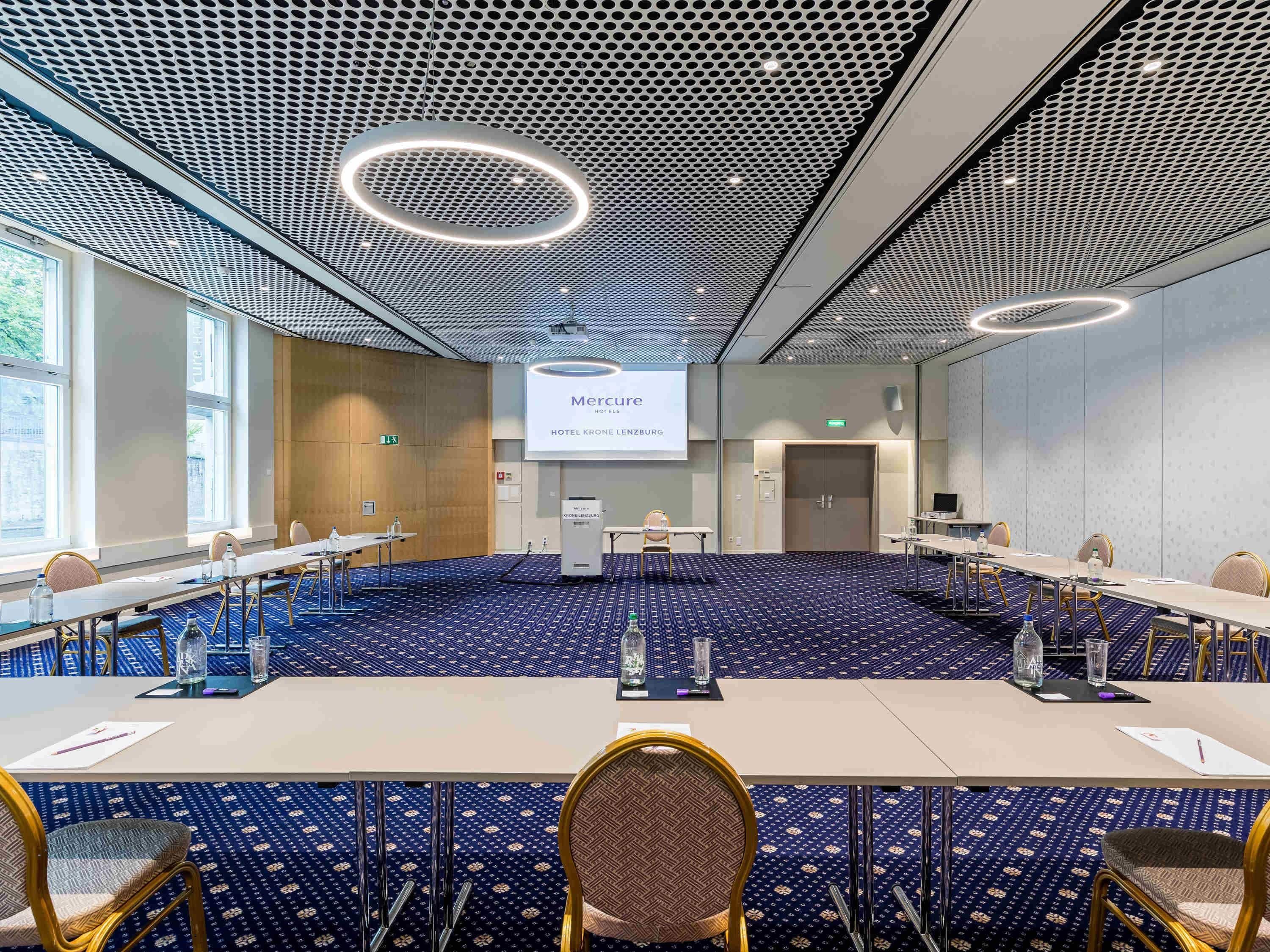 A conference room with a large screen displaying 'Mercure HOTEL KRONE LENTZBURG,' a long table with chairs, and a patterned carpet.