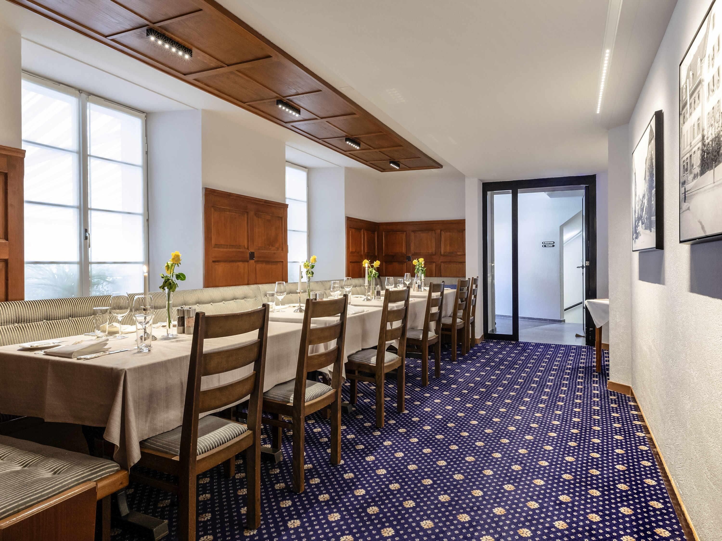 Comedor con una mesa larga dispuesta para una comida, techo de paneles de madera y alfombra estampada.