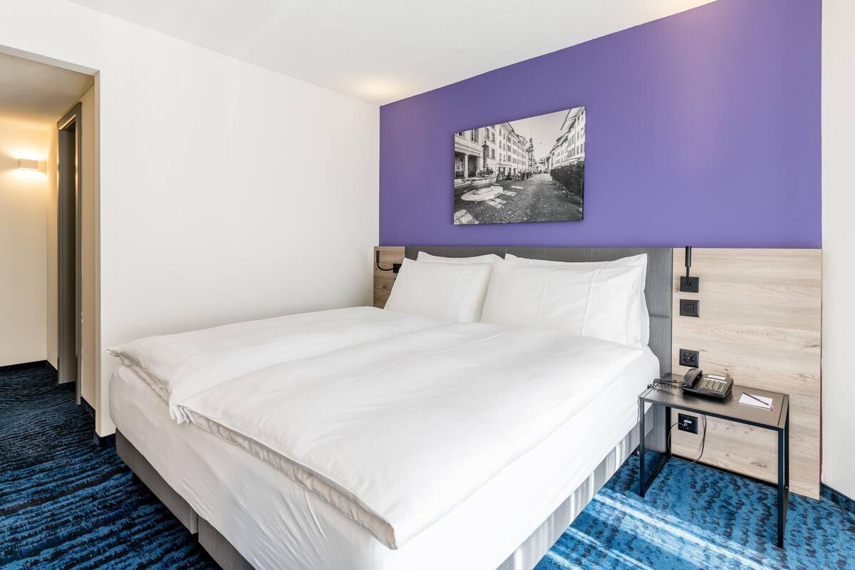 A hotel room with a bed, a nightstand with a phone, and a wall-mounted picture.