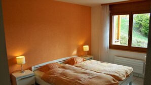 A bedroom with two beds, a nightstand with a lamp, a window with curtains, and a view of greenery outside. - Castel Club Leysin Parc Resort (Leysin)