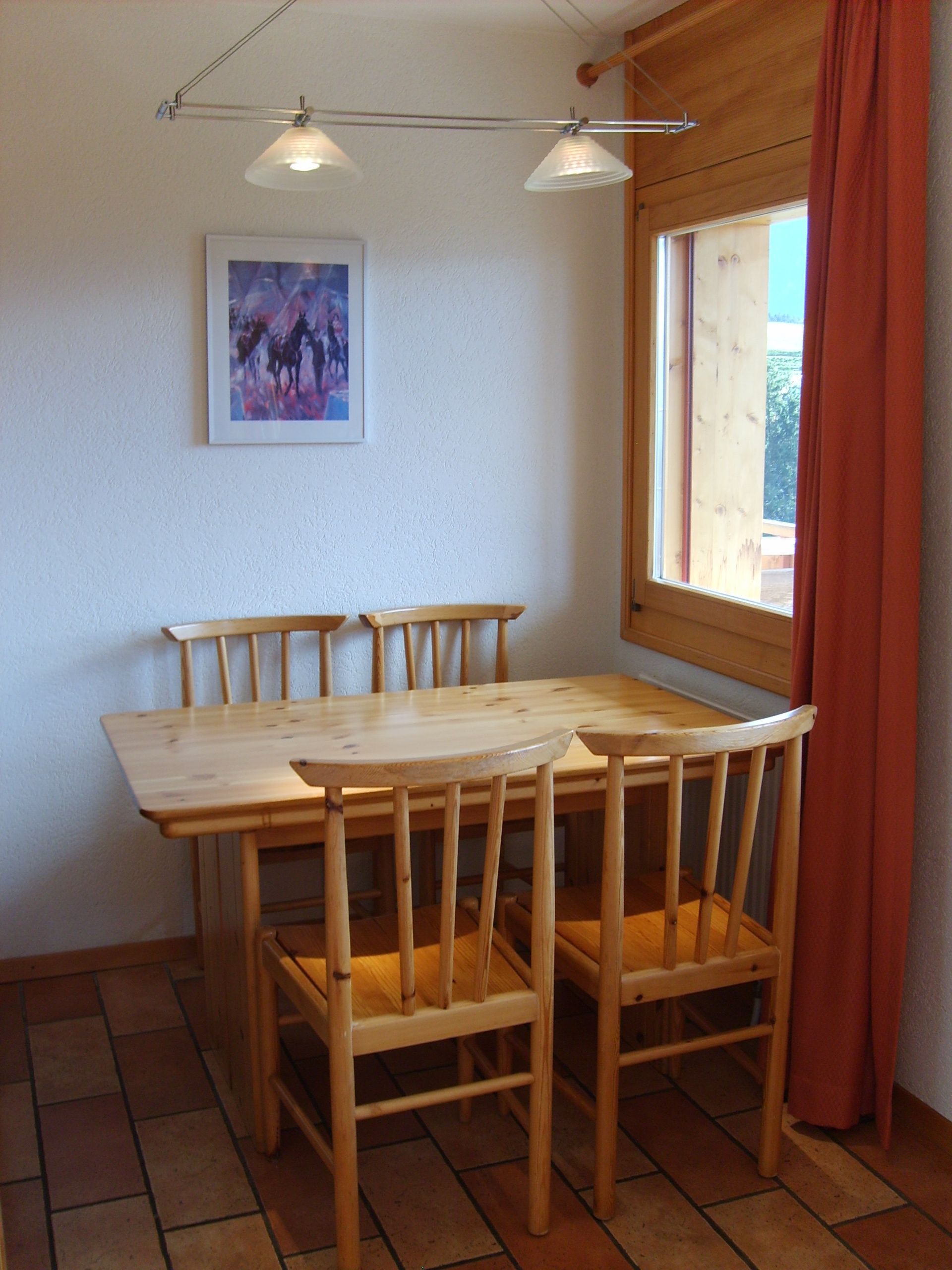 A dining area with a wooden table and chairs, a ceiling light fixture, a window with curtains, and a framed picture on the wall.