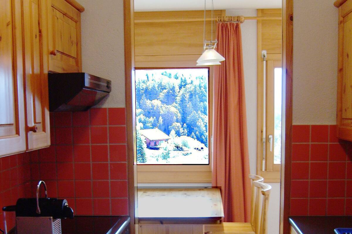 A narrow kitchen with wooden cabinets, a small table, and a window overlooking a forest.