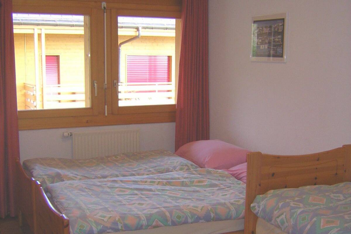 A bedroom with two wooden beds, a window with curtains, a radiator, and a small framed picture on the wall.