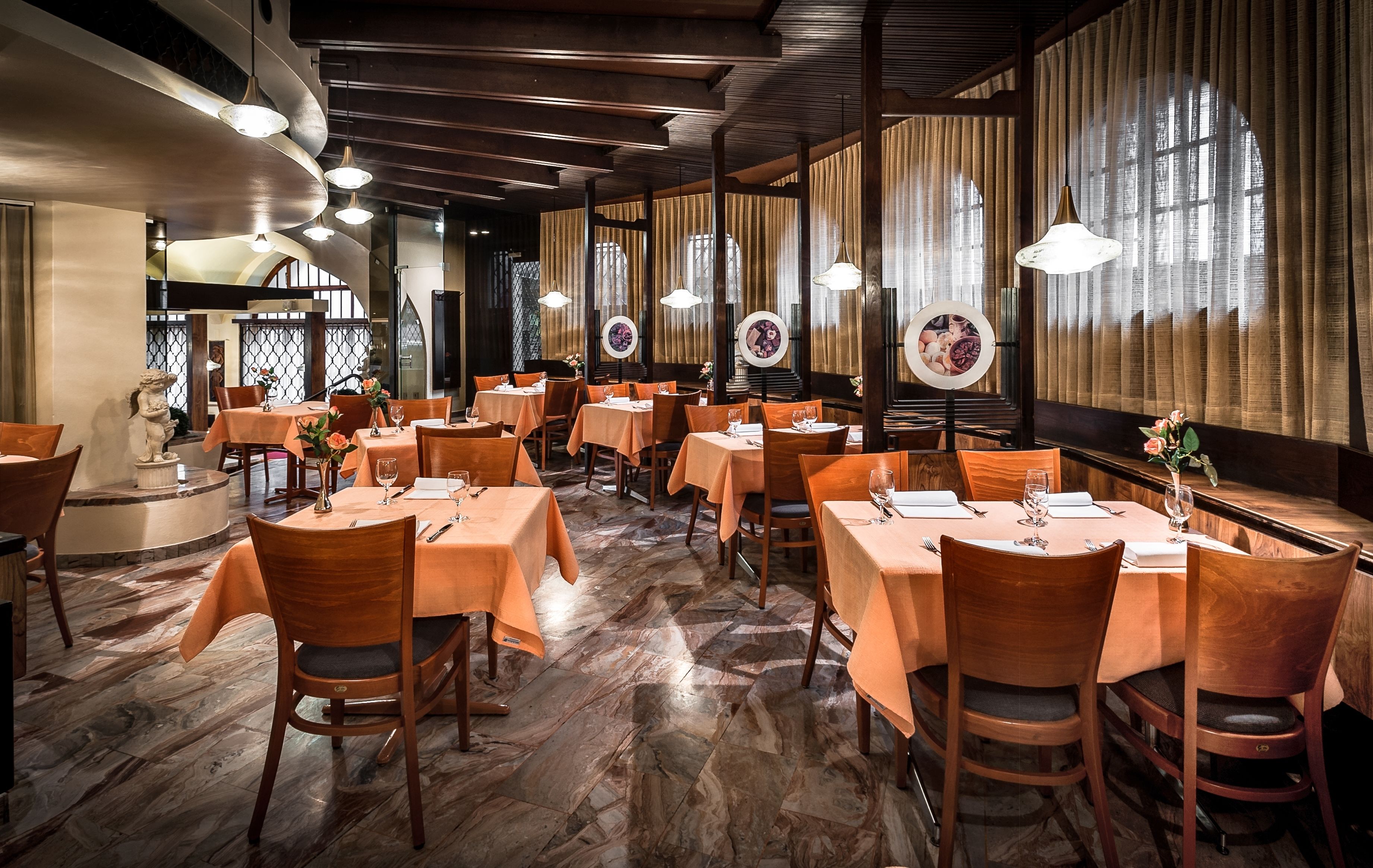 Um restaurante com várias mesas e cadeiras, piso de madeira e grandes janelas com cortinas.