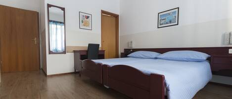 A bedroom with a wooden floor, a bed with blue bedding, a desk with a chair, a mirror, and two framed pictures on the wall.
