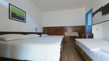 Een hotelkamer met drie enkele bedden, een houten hoofdbord, een tafeltje met tafelkleed en een ingelijst schilderij aan de muur.