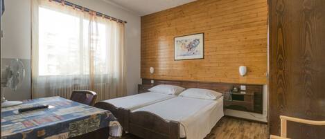 A double bed with a wooden headboard, a bedside table, a chair, a table with a quilt, and a window with curtains.