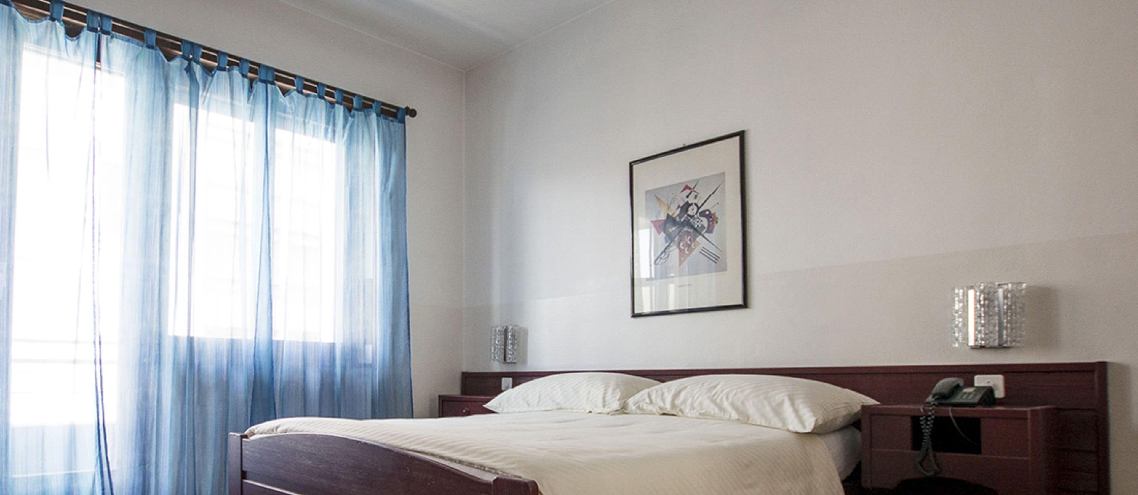 Ein Hotelzimmer mit einem Bett, einem Telefon, einem gerahmten Bild an der Wand und einem Fenster mit Vorhängen.