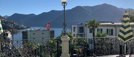A terrace with a view of mountains and a lake, featuring a lamp post, a table, and chairs.