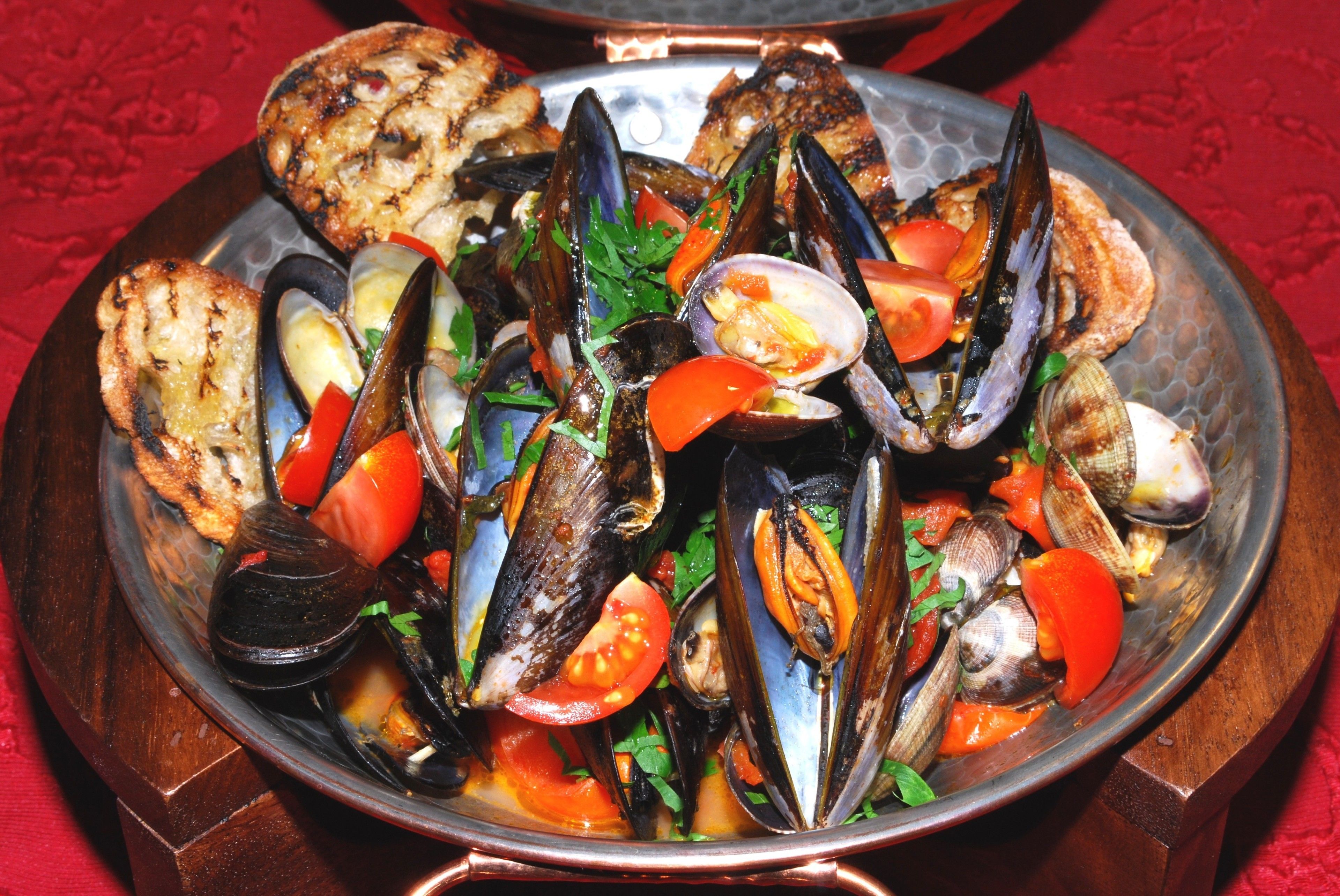 A seafood dish with mussels, tomatoes, and herbs, served on a metal plate with a red tablecloth.
