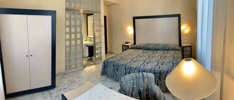 A hotel room with a bed, bedside tables, a desk, a chair, a wardrobe, and a bathroom visible through a partition.
