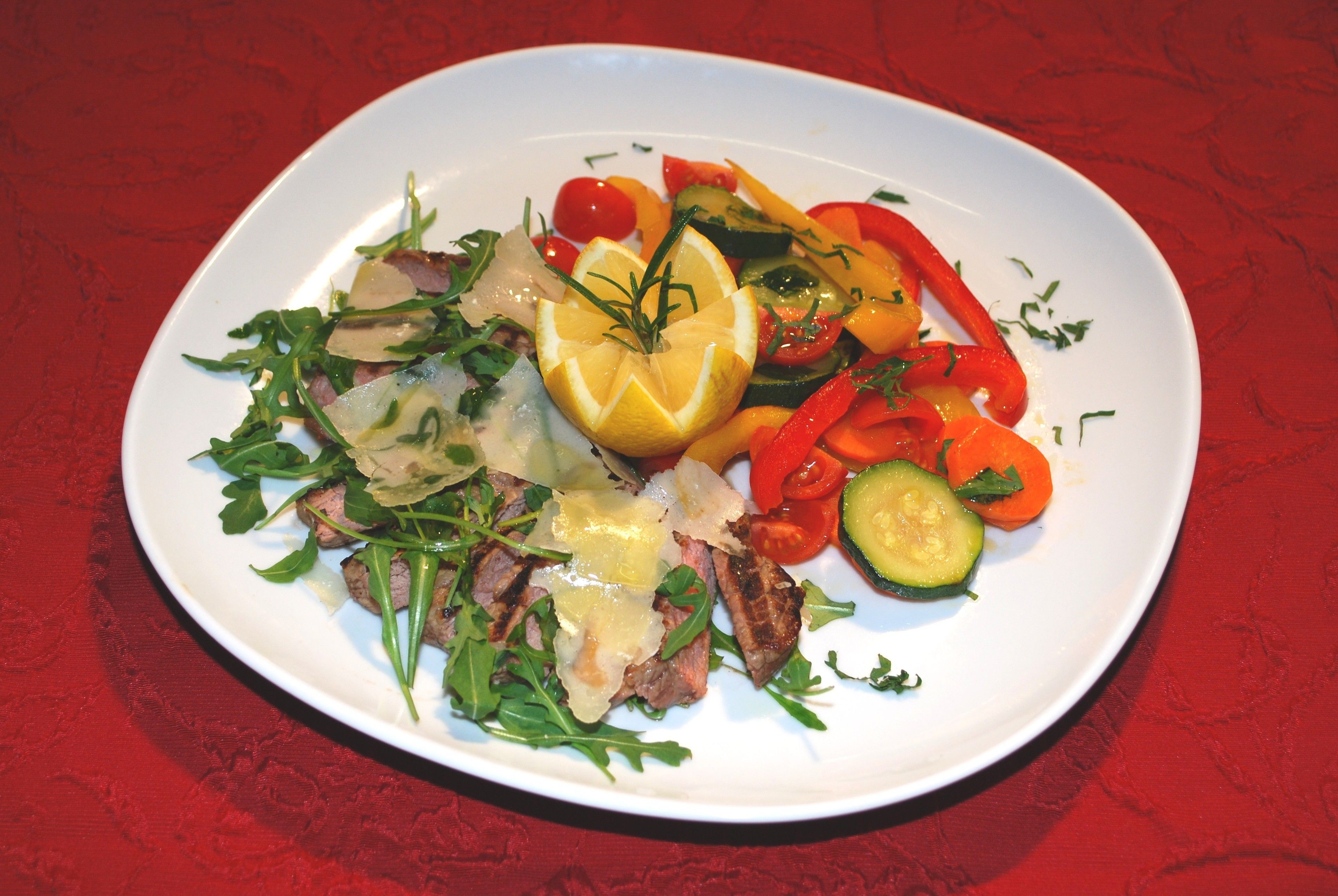 A plate of food with grilled meat, vegetables, and a lemon wedge on a red tablecloth.