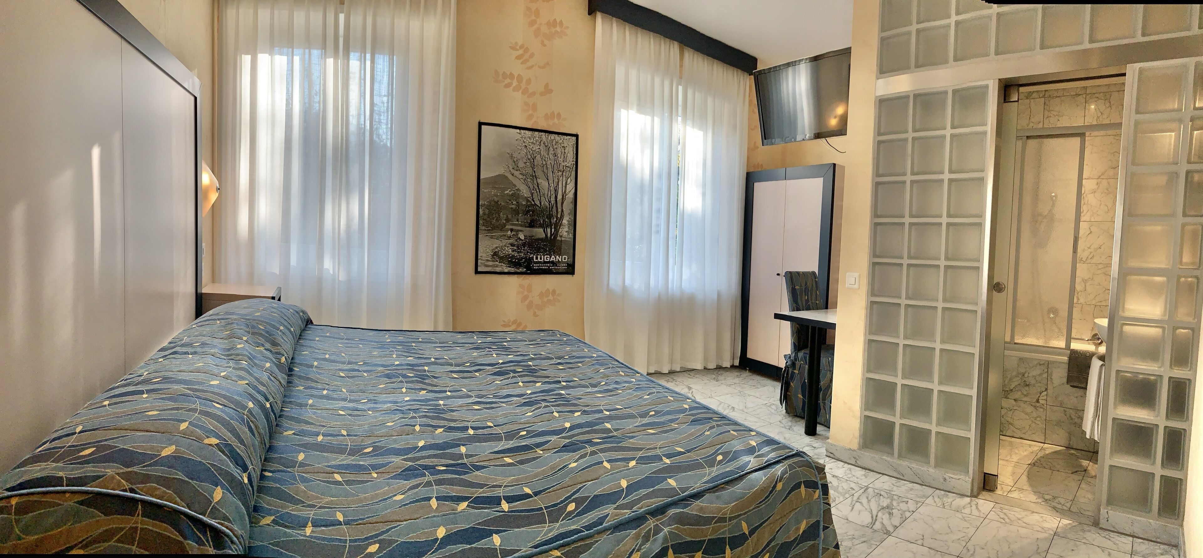 Un dormitorio con cama, escritorio, televisor y baño con puerta de vidrio.