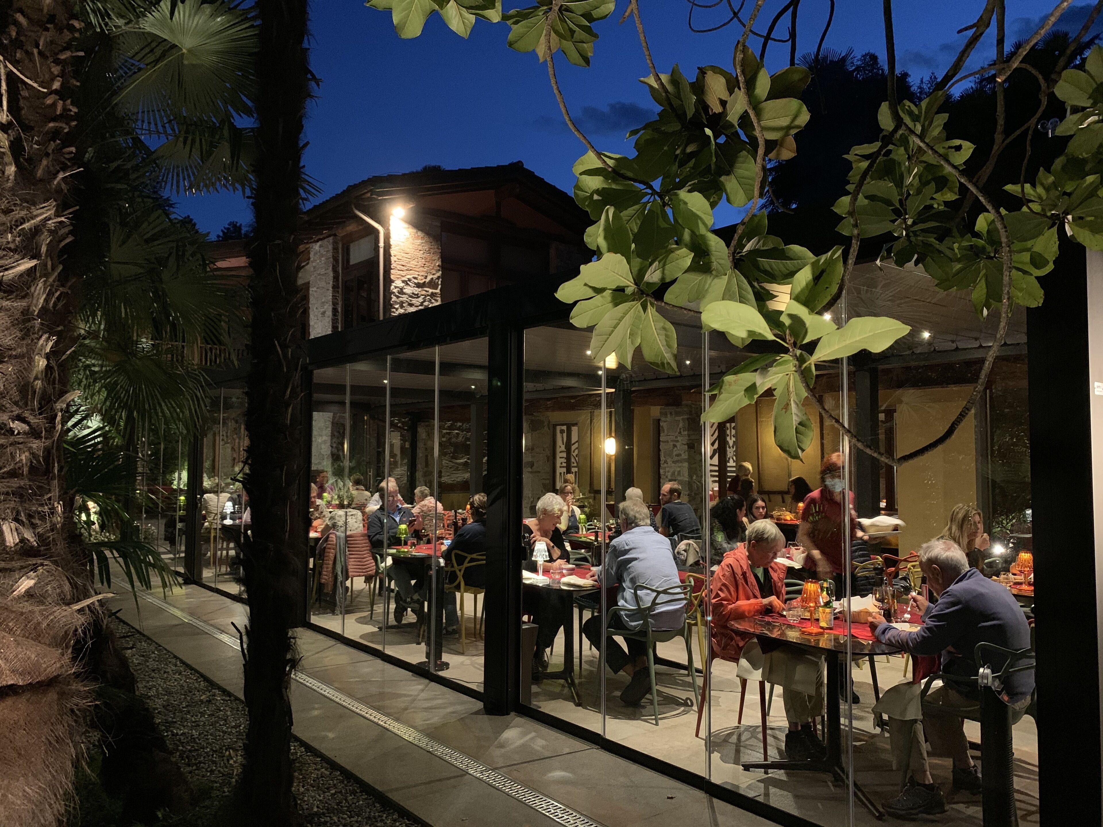 An outdoor dining area with glass walls, surrounded by trees and a stone building.