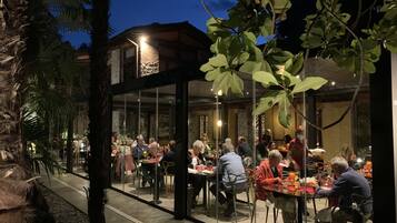 An outdoor dining area with glass walls, surrounded by trees and a stone building.