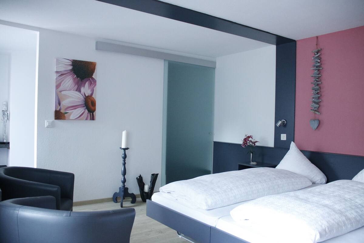 A modern bedroom with a bed, a chair, a painting, and a bedside table with a vase.
