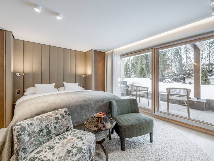 Un dormitorio moderno con una cama grande, una pequeña mesa redonda con un postre, un sillón verde y vistas al exterior a través de amplias ventanas.