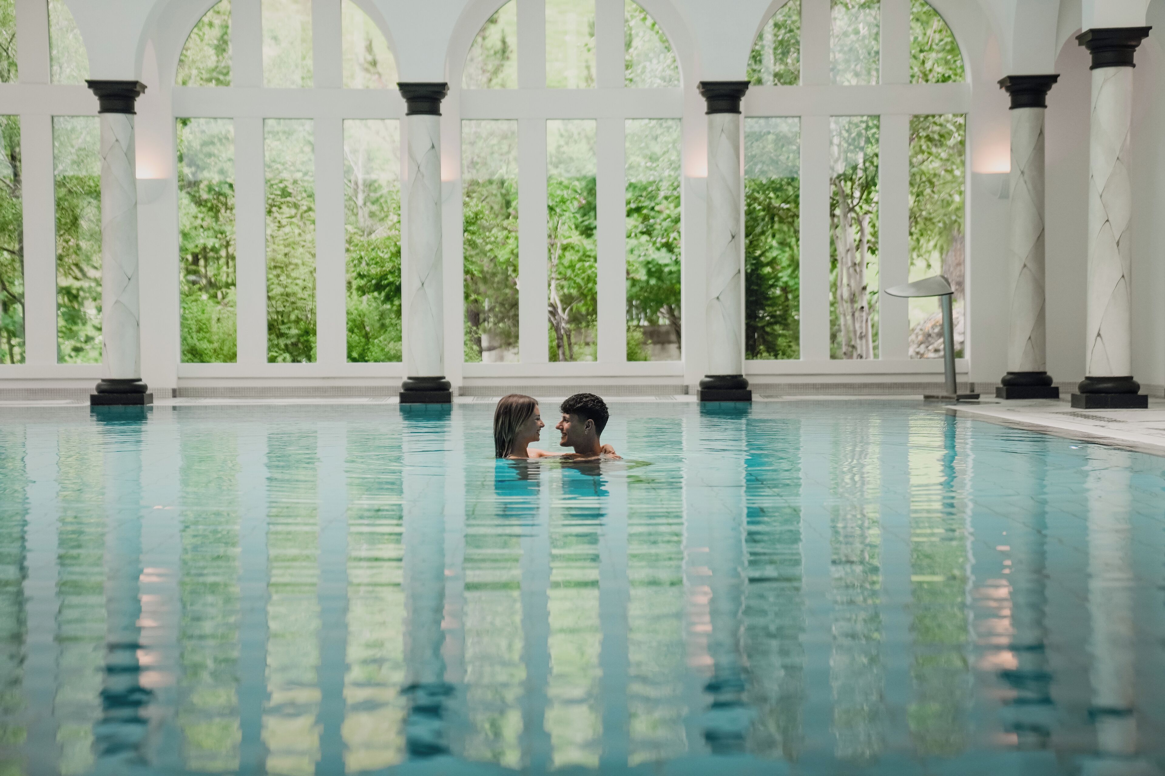 Piscina interna con pareti di vetro trasparenti, colonne e vista sul verde circostante.