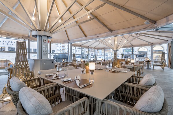 A spacious dining area with high ceilings and large windows, featuring wicker furniture and a central table set for dining. - Relais & Châteaux Chasa Montana (Samnaun)