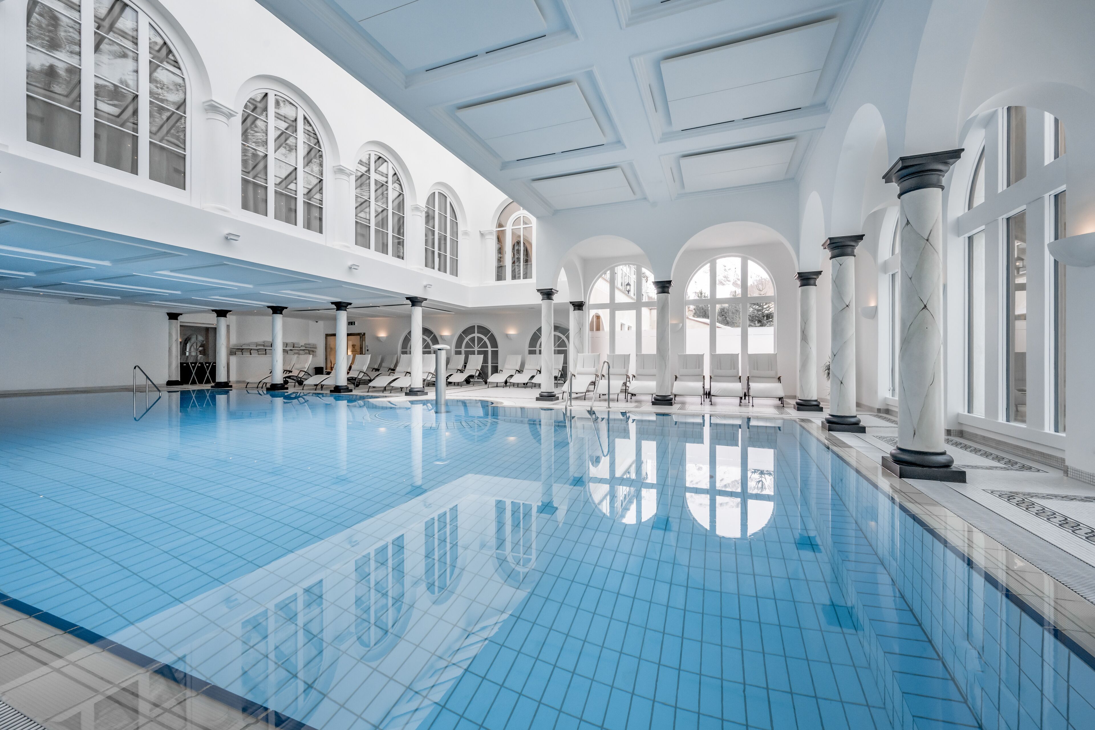 A large indoor swimming pool with clear blue tiles, white columns, and arched windows.