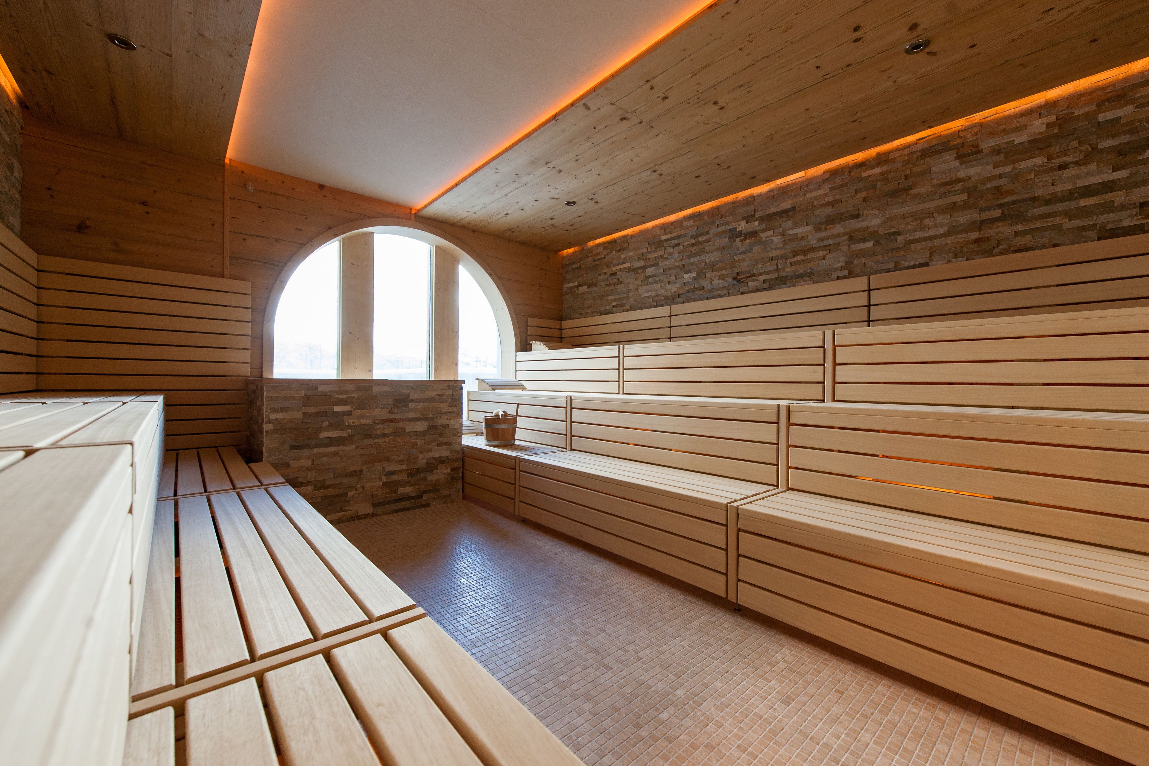 A sauna with wooden benches and a stone wall, a large arched window, and a wooden ceiling.