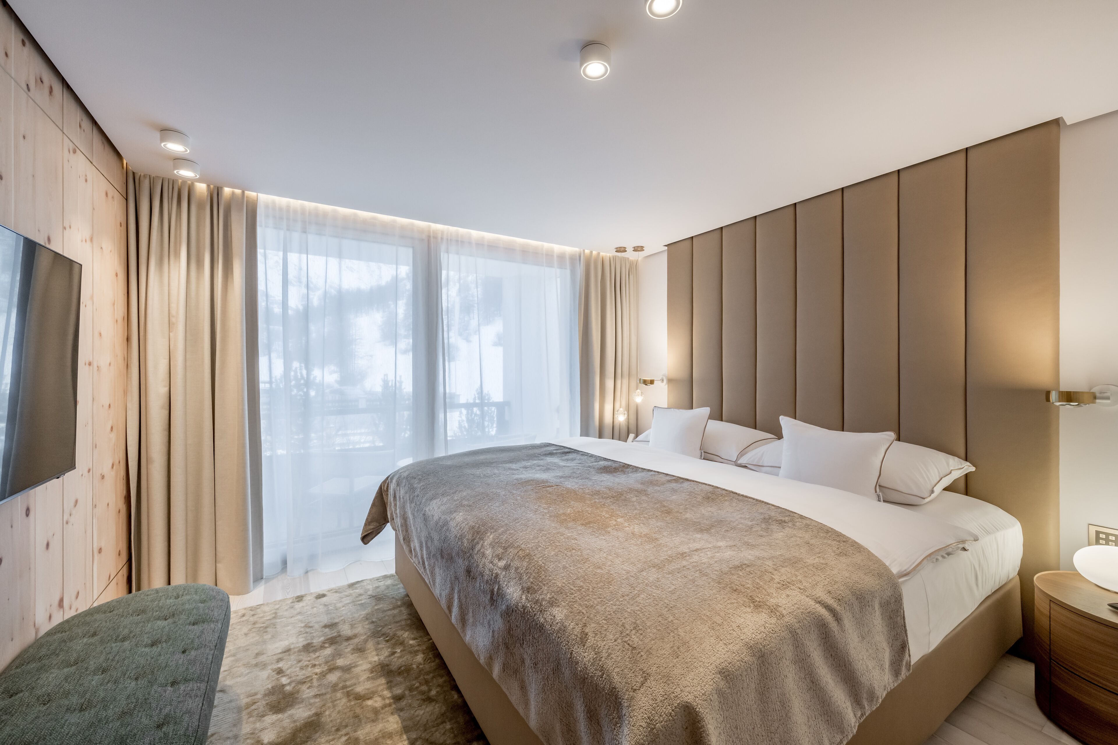 Une chambre moderne avec un grand lit, un mur d’accent en bois, une télévision et une grande fenêtre avec des rideaux.
