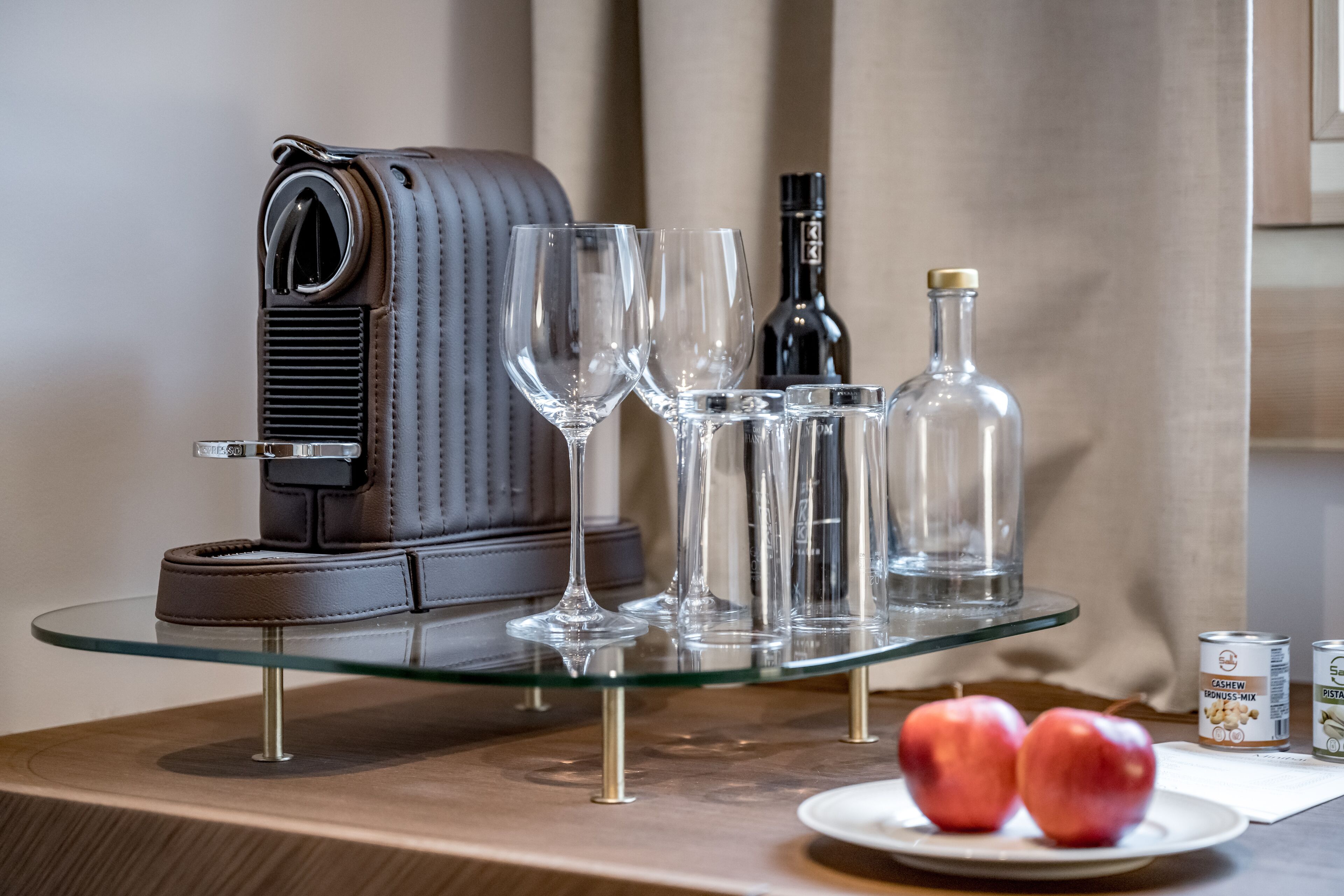 A glass table with a coffee maker, wine glasses, bottles, and apples.